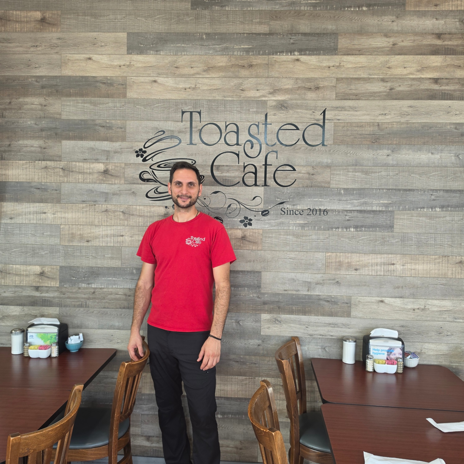 Owner of Toasted Cafe standing in front of the Toasted Cafe wall logo