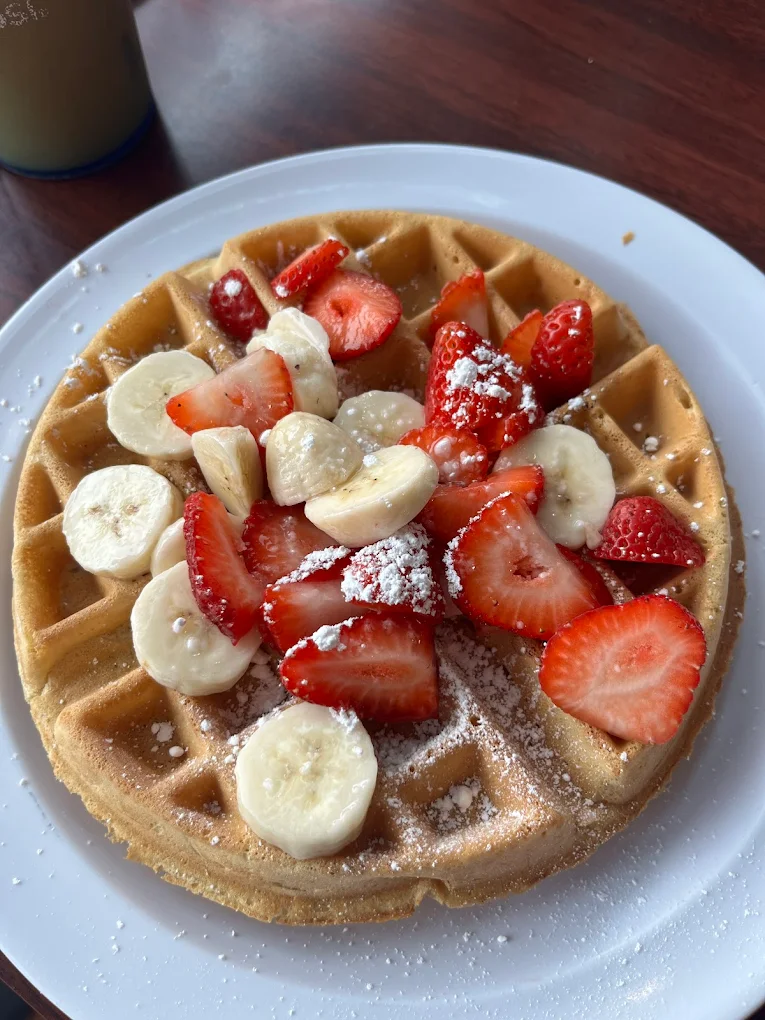 Belgian Waffle & Fresh Fruit at Toasted Cafe — Crisp golden waffle piled high with sliced strawberries, banana and a dusting of powdered sugar.